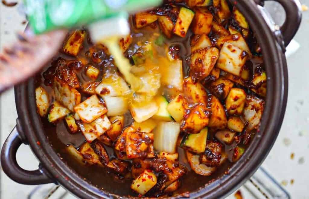 A pot filled with chopped vegetables, including zucchini and onions, mixed with doenjang. A yellow liquid is being poured over the vegetables, creating a vibrant, colorful dish.